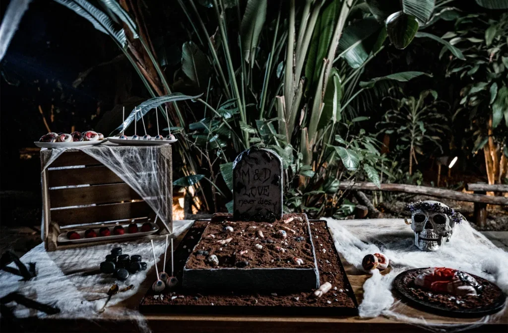 Ein dekorativer Sweet Table im Gothic-Stil mit Grabstein, Totenkopf und Spinnweben. Umgeben von üppigem, tropischem Pflanzenwerk, ideal für eine düstere Hochzeit.