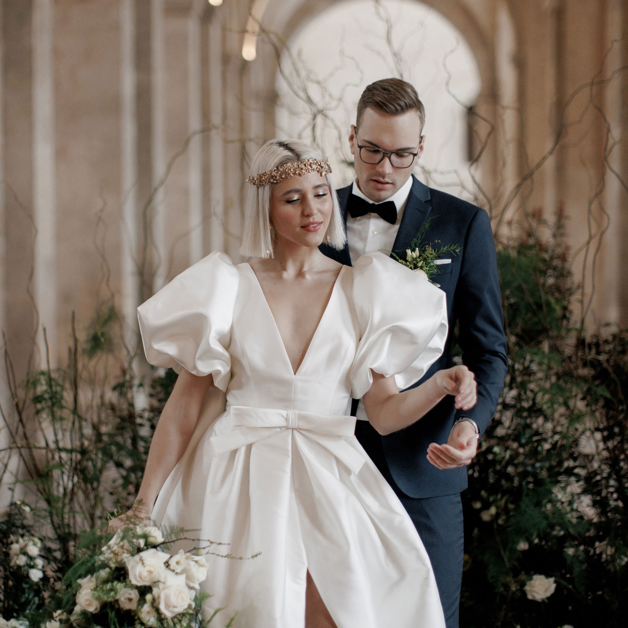 Das Brautpaar steht in einem festlichen Saal, die Braut beeindruckt mit einem ausgefallenen Hochzeitskleid mit Puffärmeln, während der Bräutigam einen eleganten Anzug trägt.