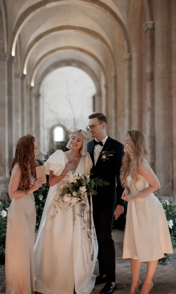 Das Brautpaar steht lächelnd mit zwei Brautjungfern unter den Bögen eines historischen Korridors. Die Braut, in einem eleganten weißen Kleid mit Krone, und der Bräutigam, in einem Anzug, genießen einen freudigen Moment mit ihren Begleiterinnen.