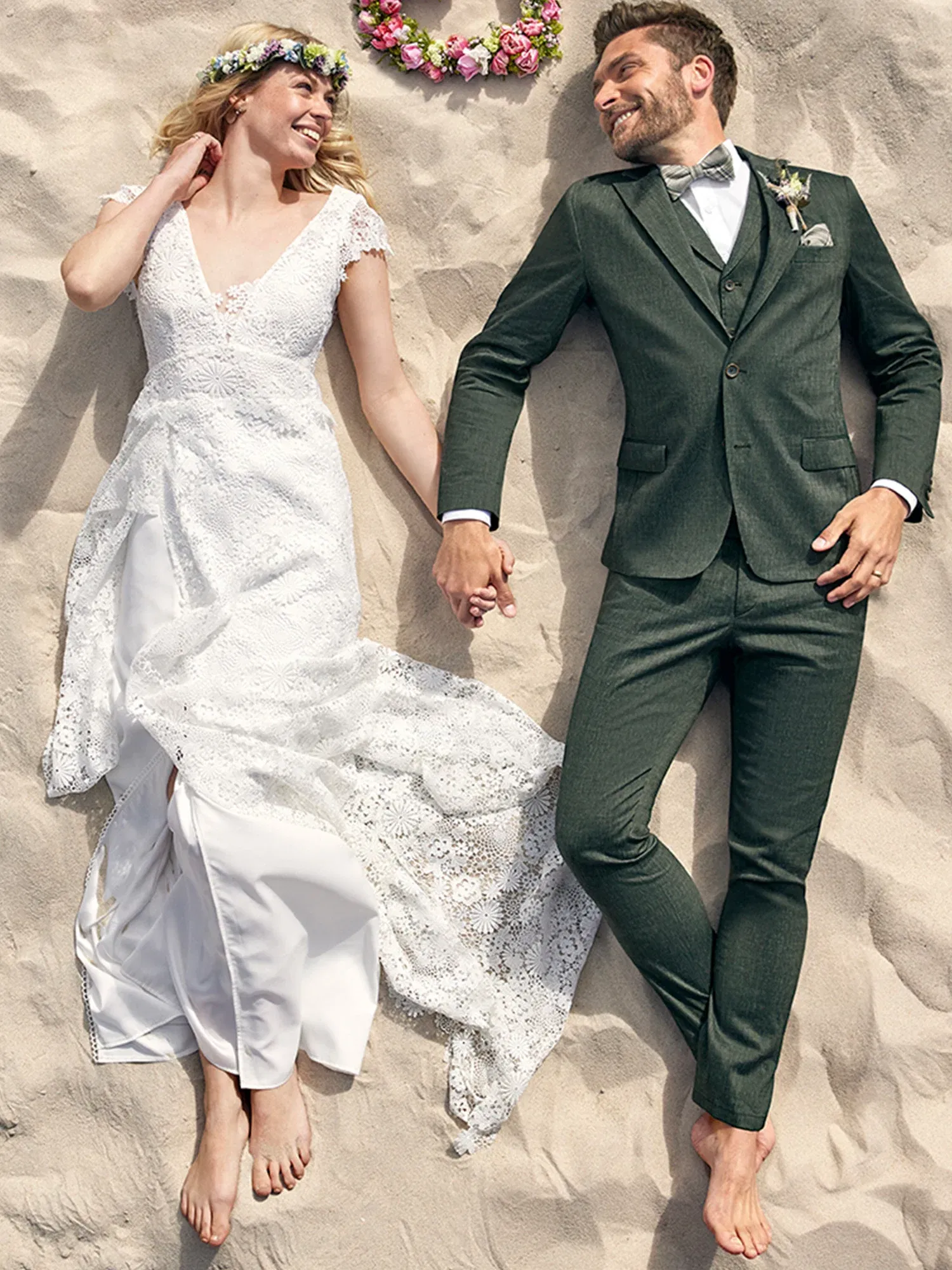 Ein frisch verheiratetes Paar liegt lächelnd im Sand. Die Braut in einem Spitzenhochzeitskleid und der Bräutigam in einem grünen Anzug schauen einander verliebt an und halten sich an den Händen.
