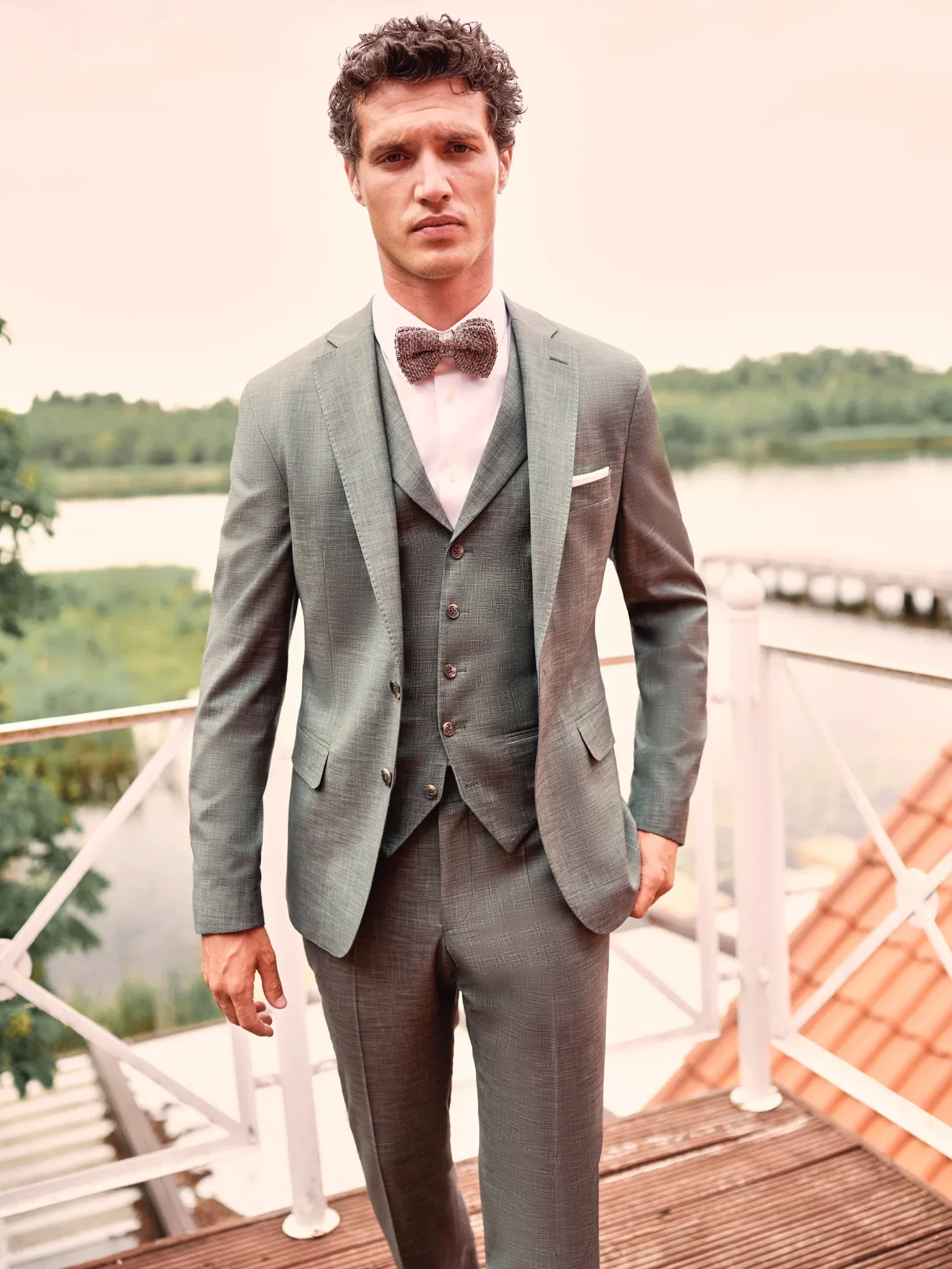 Eleganter Mann in einem graugrünen Anzug mit Weste und Fliege, der auf einem Balkon vor einem idyllischen Seepanorama steht.