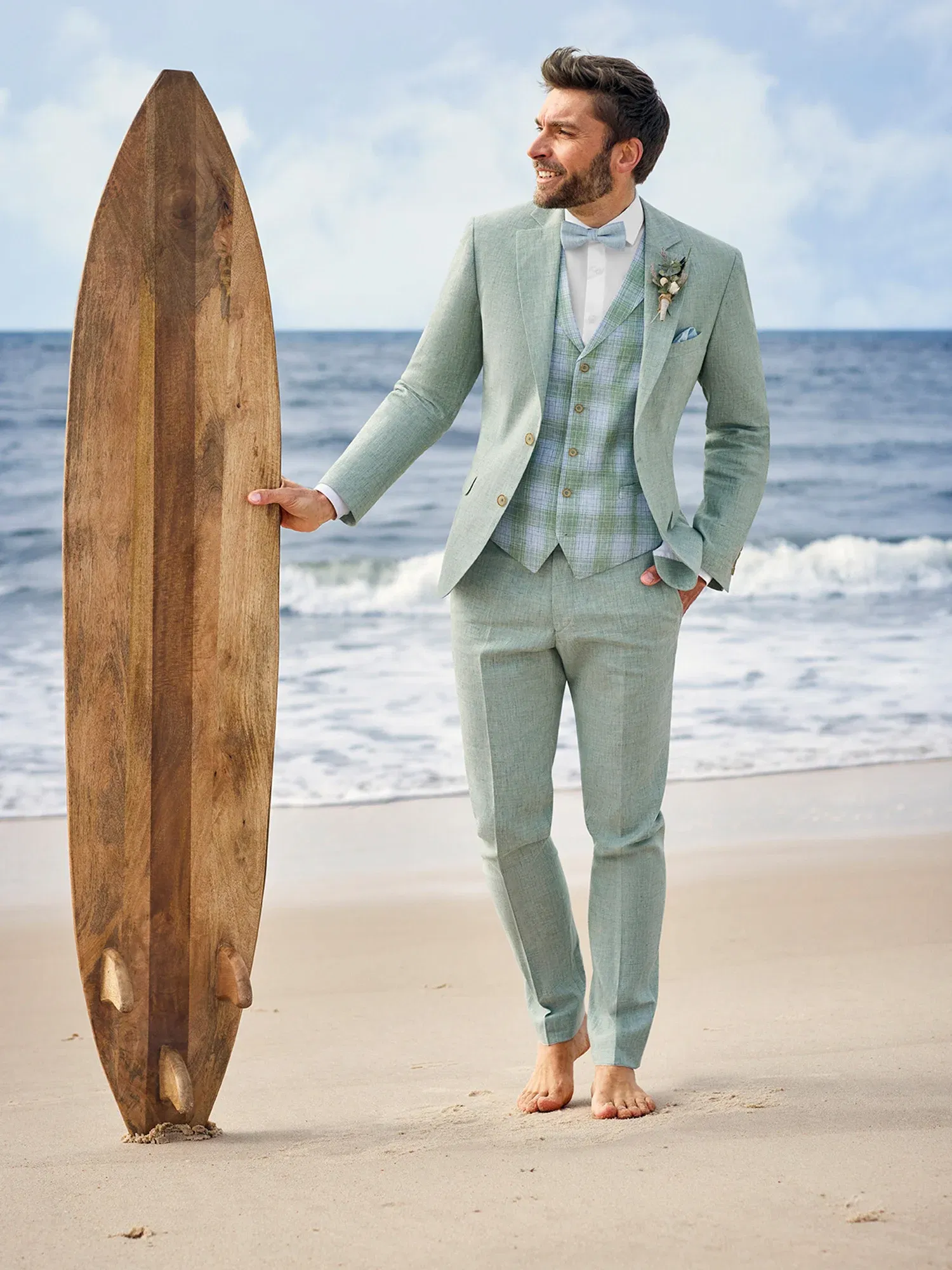 Eleganter Bräutigam in einem mintfarbenen Anzug und Fliege steht am Strand mit einem Holz-Surfbrett, barfuß und mit Blick auf das Meer.