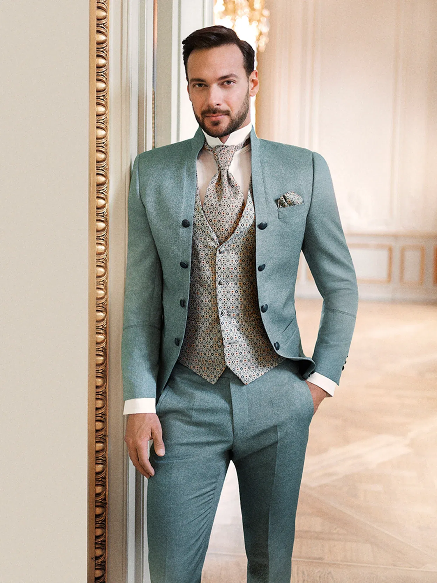 Ein Bräutigam in einem festlichen, grünen Anzug mit dekorativem Plastron und gemusterter Weste lehnt entspannt an einer goldgerahmten Wand in einem opulenten Raum.