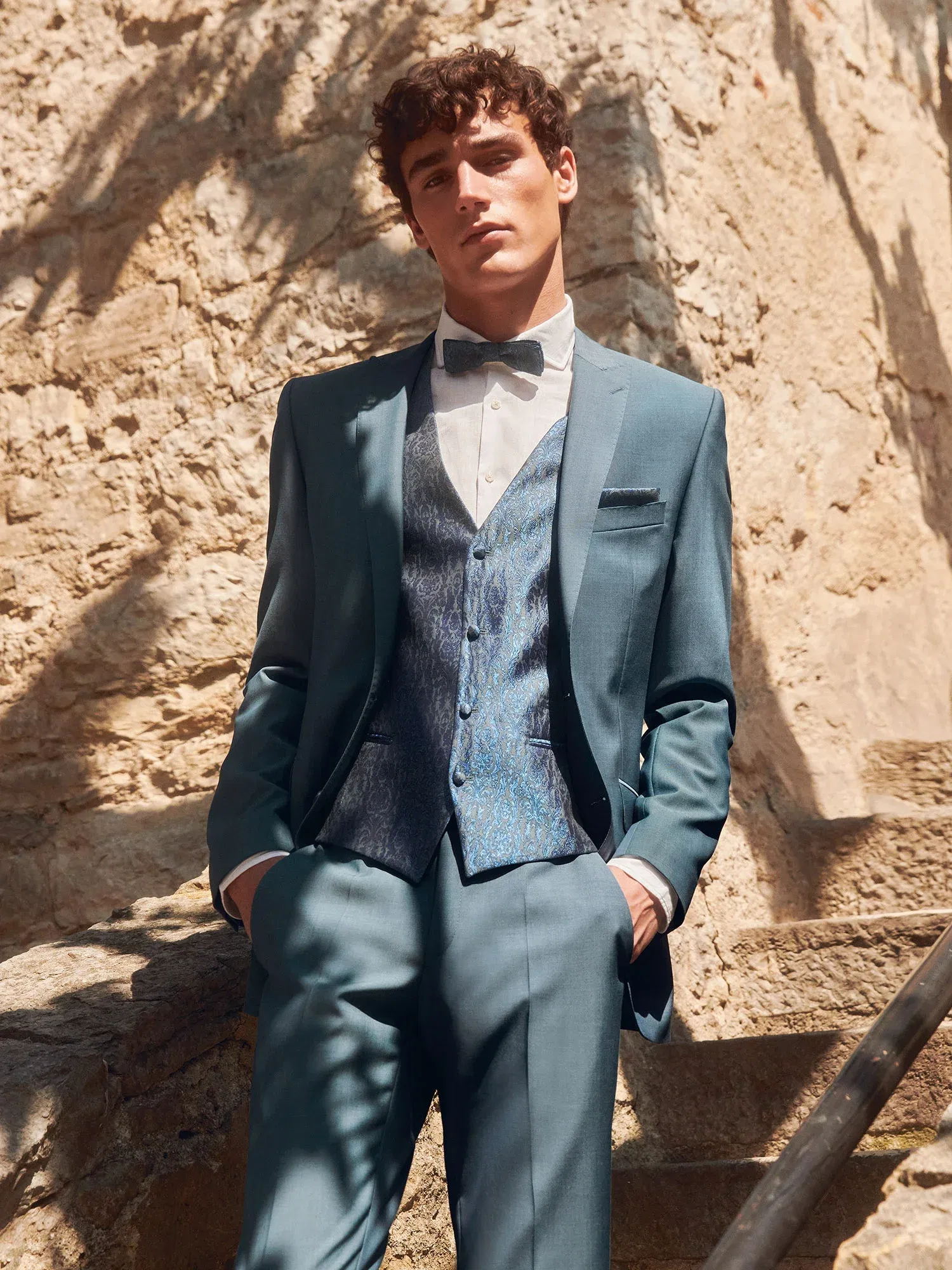 Stilvoller Mann in einem mintfarbenen Anzug und einer schimmernden Weste, der lässig vor einer rustikalen Steinmauer steht und in die Kamera blickt.