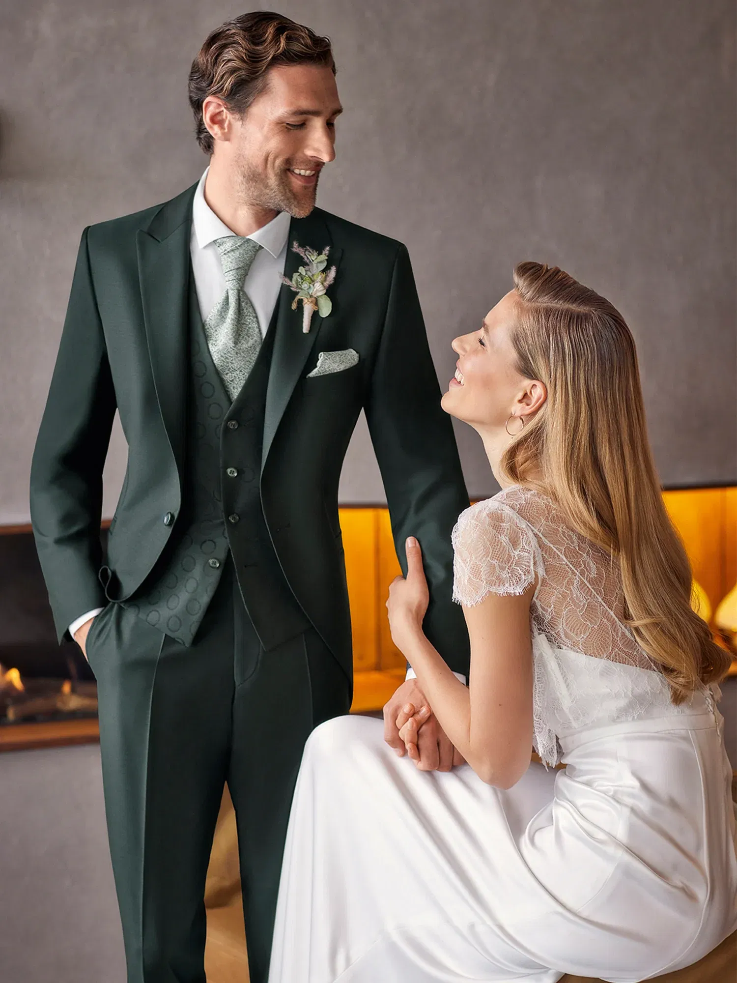 Ein Bräutigam in einem dunkelgrünen Hochzeitsanzug steht neben seiner Braut, die ihn bewundernd ansieht, während sie sich sanft an seine Hand lehnt.