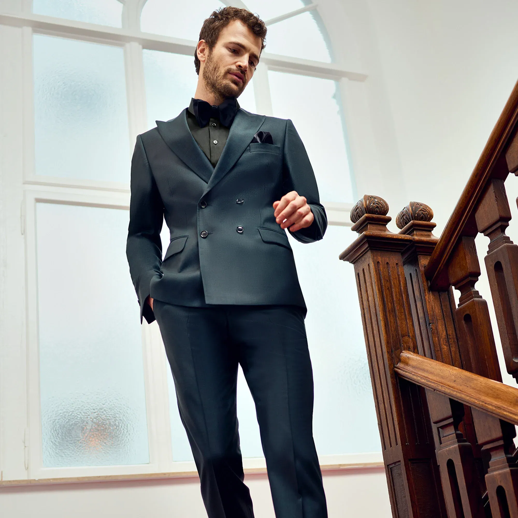 Bräutigam in einem edlen, zweireihigen Anzug in dunklen Tönen, der selbstbewusst eine Holztreppe in einem historischen Gebäude hinunterschreitet.