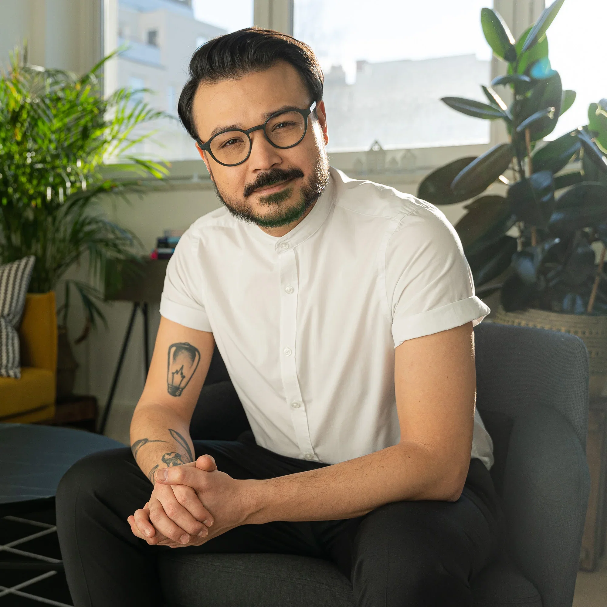 Psychotherapeut Umut C. Özdemir sitzt entspannt auf einem Sessel in einem modern eingerichteten Raum und lächelt in die Kamera, umgeben von Pflanzen.