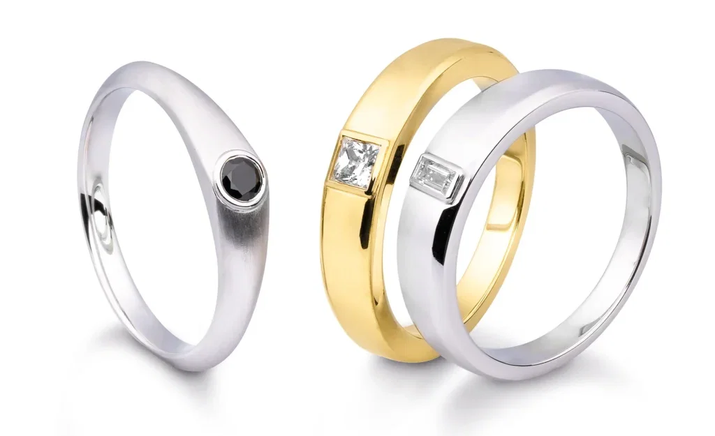 Eine Auswahl von drei Eheringen: ein schlichter Silberring mit schwarzem Stein und zwei kombinierte Ringe aus Gold und Silber mit Diamanten.