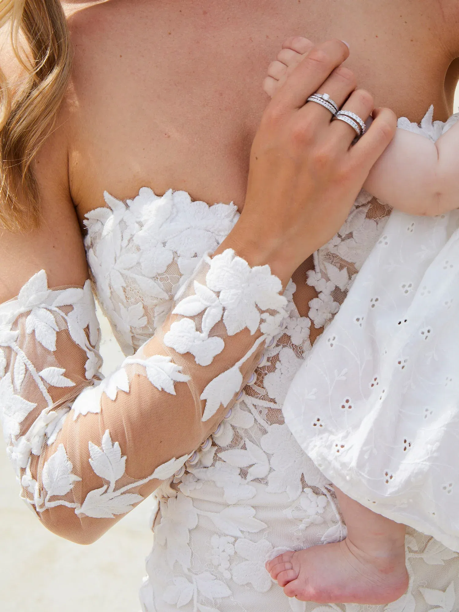 Eine Braut in einem Spitzenkleid hält ihr Baby. Der Fokus liegt auf den Blumenverzierungen des Kleides und den Eheringen, während das Baby ihre Hand greift.
