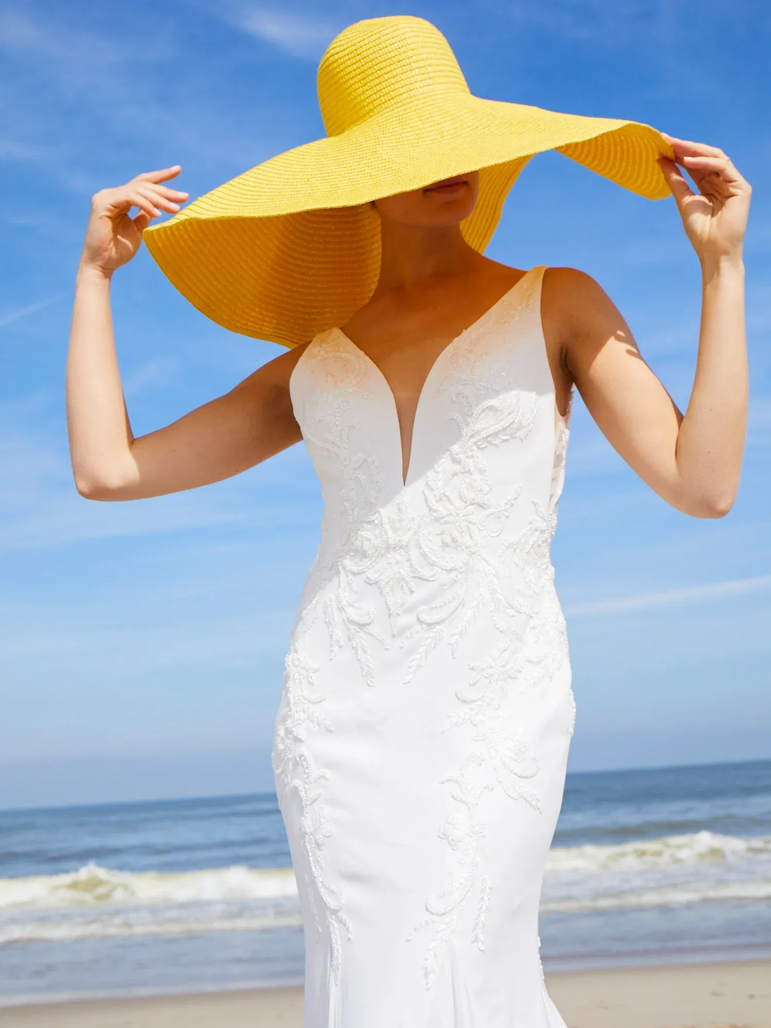 Eine Braut in einem weißen, figurbetonten Hochzeitskleid mit filigranen Stickereien hält einen breiten, gelben Sonnenhut am Strand.