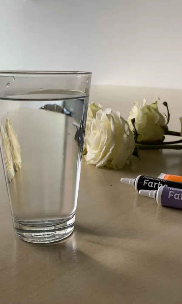 Ein schlichtes Glas Wasser steht auf einem Tisch, umgeben von weißen Rosen. Drei Tuben Lebensmittelfarbe in Lila, Schwarz und Orange liegen im Hintergrund, bereit für ein Färbeprojekt.