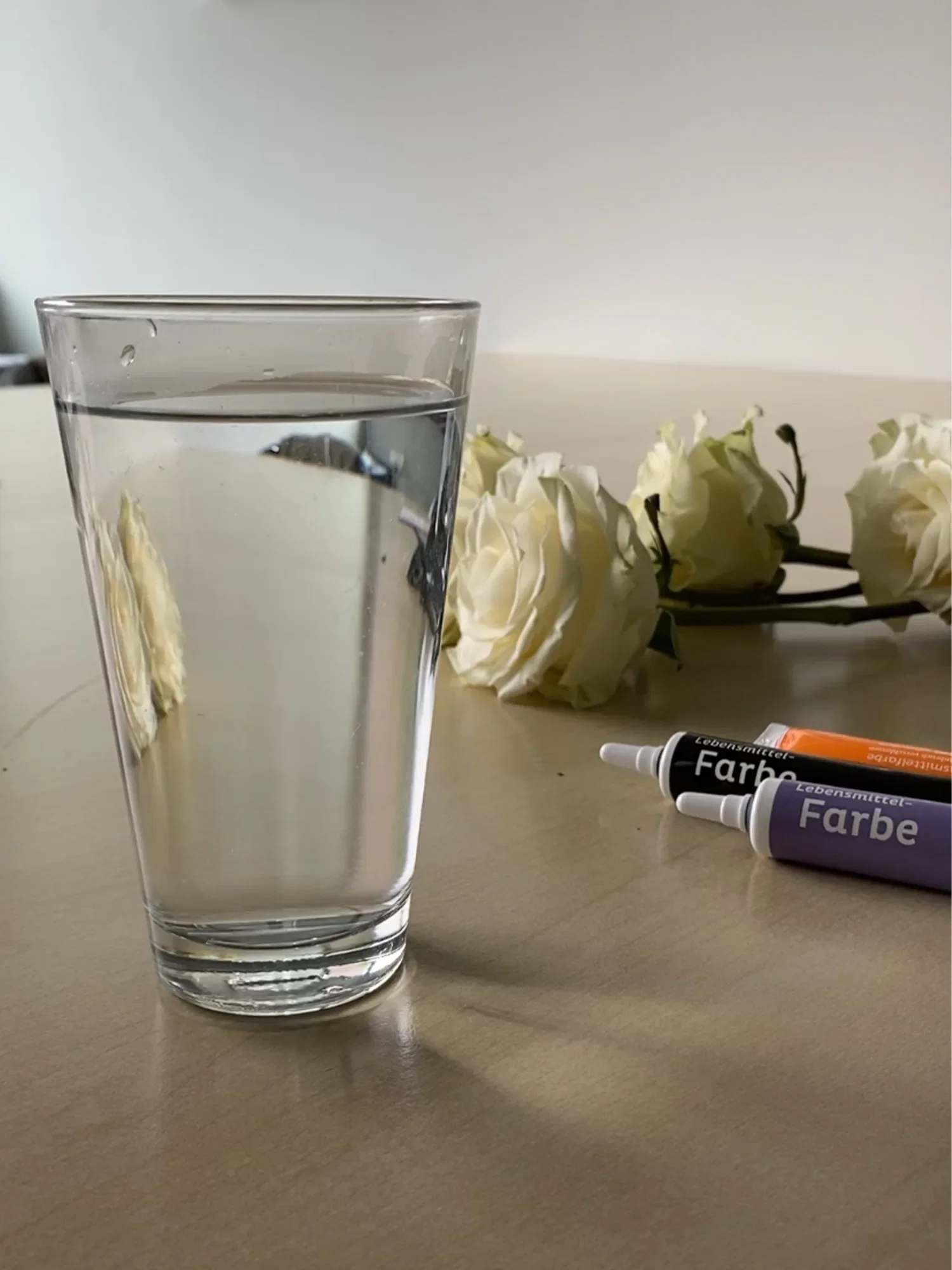 Ein schlichtes Glas Wasser steht auf einem Tisch, umgeben von weißen Rosen. Drei Tuben Lebensmittelfarbe in Lila, Schwarz und Orange liegen im Hintergrund, bereit für ein Färbeprojekt.