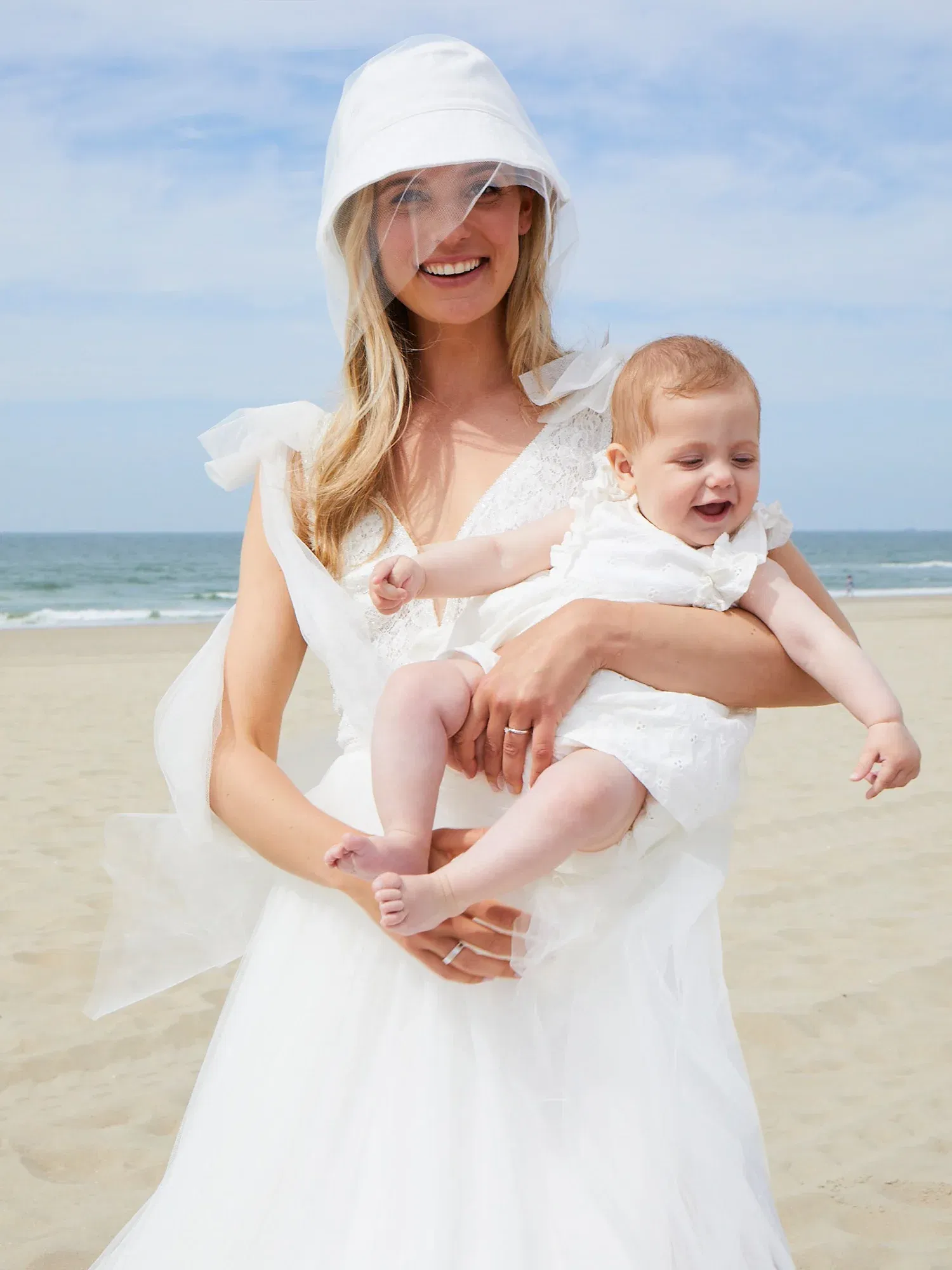 Eine Braut in einem eleganten Hochzeitskleid und Hut hält lächelnd ihr Baby am Strand. Beide sind in strahlend weißen Outfits gekleidet.
