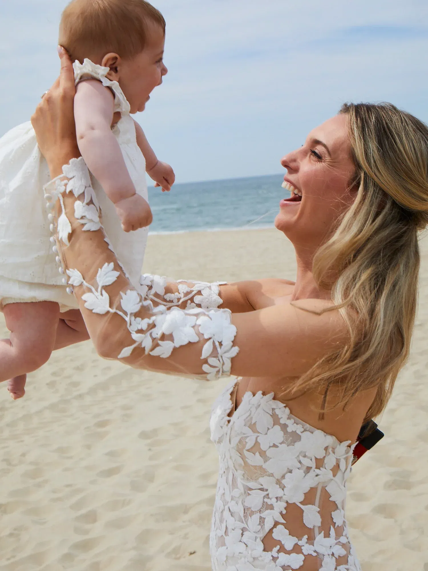 Eine Braut am Strand, die ihr Baby lachend hochhebt. Sie trägt ein florales Spitzenkleid, und die Szene vermittelt pure Freude.