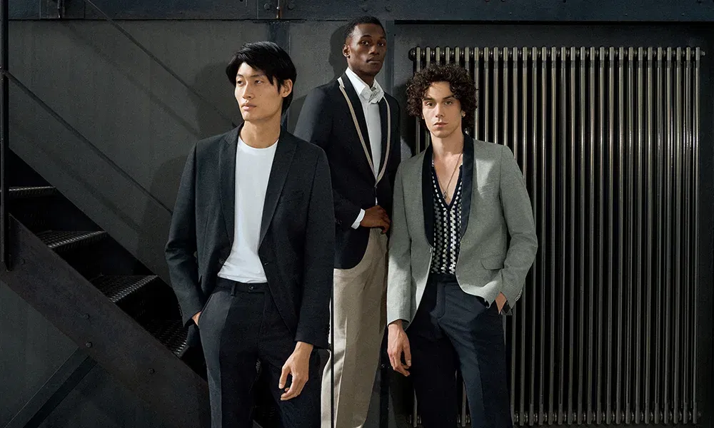 Drei stilvolle Männer posieren in einer modernen, industriellen Umgebung. Links trägt ein Mann einen klassischen schwarzen Anzug kombiniert mit einem schlichten weißen T-Shirt. Der Mann in der Mitte wählt einen lässigen Look mit einem dunkelblauen Blazer und beigen Hosen. Der dritte Mann rechts vervollständigt das Trio mit einem grauen Blazer und einem auffälligen gemusterten Hemd.