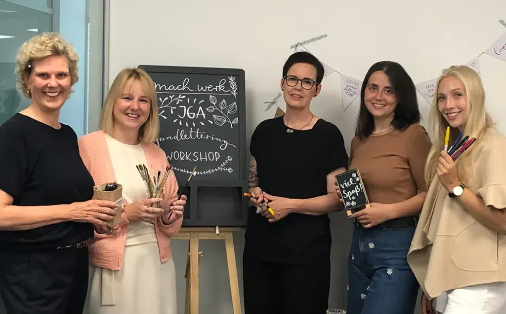 Fünf Frauen nehmen an einem Handlettering-Kurs teil und lächeln gemeinsam für ein Gruppenfoto. Einige von ihnen halten Stifte, während eine Tafel im Hintergrund die Arbeit des Workshops zeigt.
