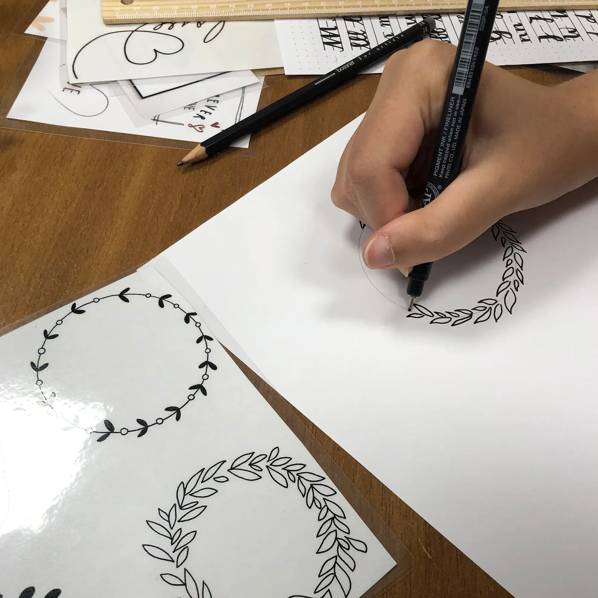 Eine Hand zeichnet einen dekorativen Blätterkranz auf ein weißes Papier. Neben der Zeichnung liegen Vorlagen mit verschiedenen Kranzmustern und ein Bleistift auf dem Tisch.