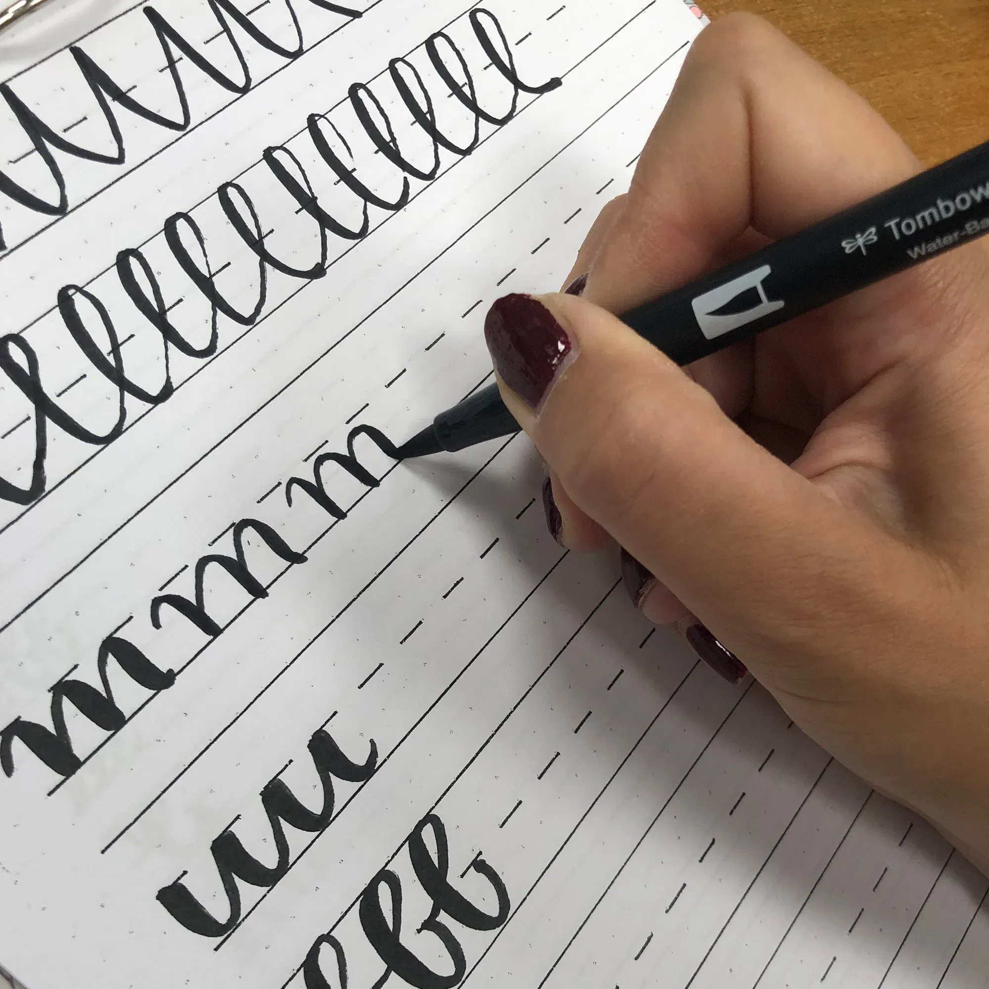Eine Person übt mit einem Tombow-Pinselstift Handlettering auf einem Blatt mit vorgegebenen Linien. Die Hand zeigt sorgfältige Striche und elegante Buchstabenformen auf dem Übungsblatt.