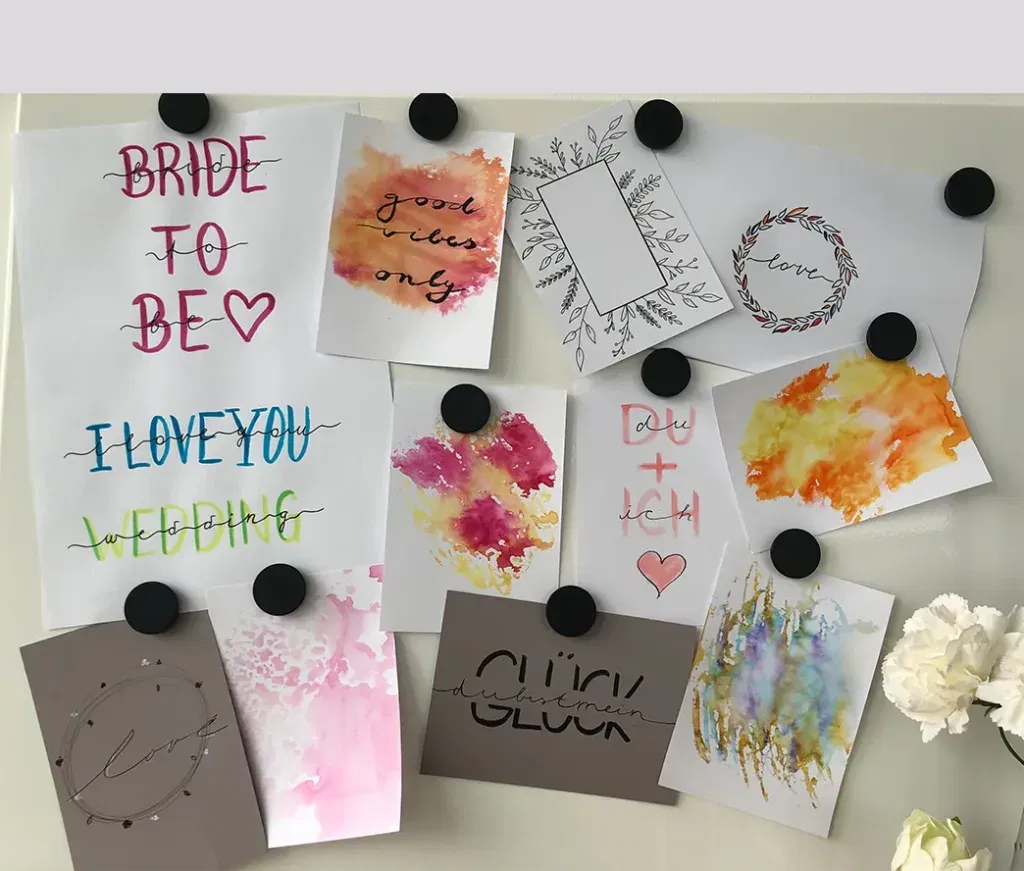 Eine Auswahl von Handlettering-Arbeiten, die an einer Pinnwand befestigt sind, zeigen bunte Schriftzüge und Aquarell-Motive für Hochzeitsthemen.