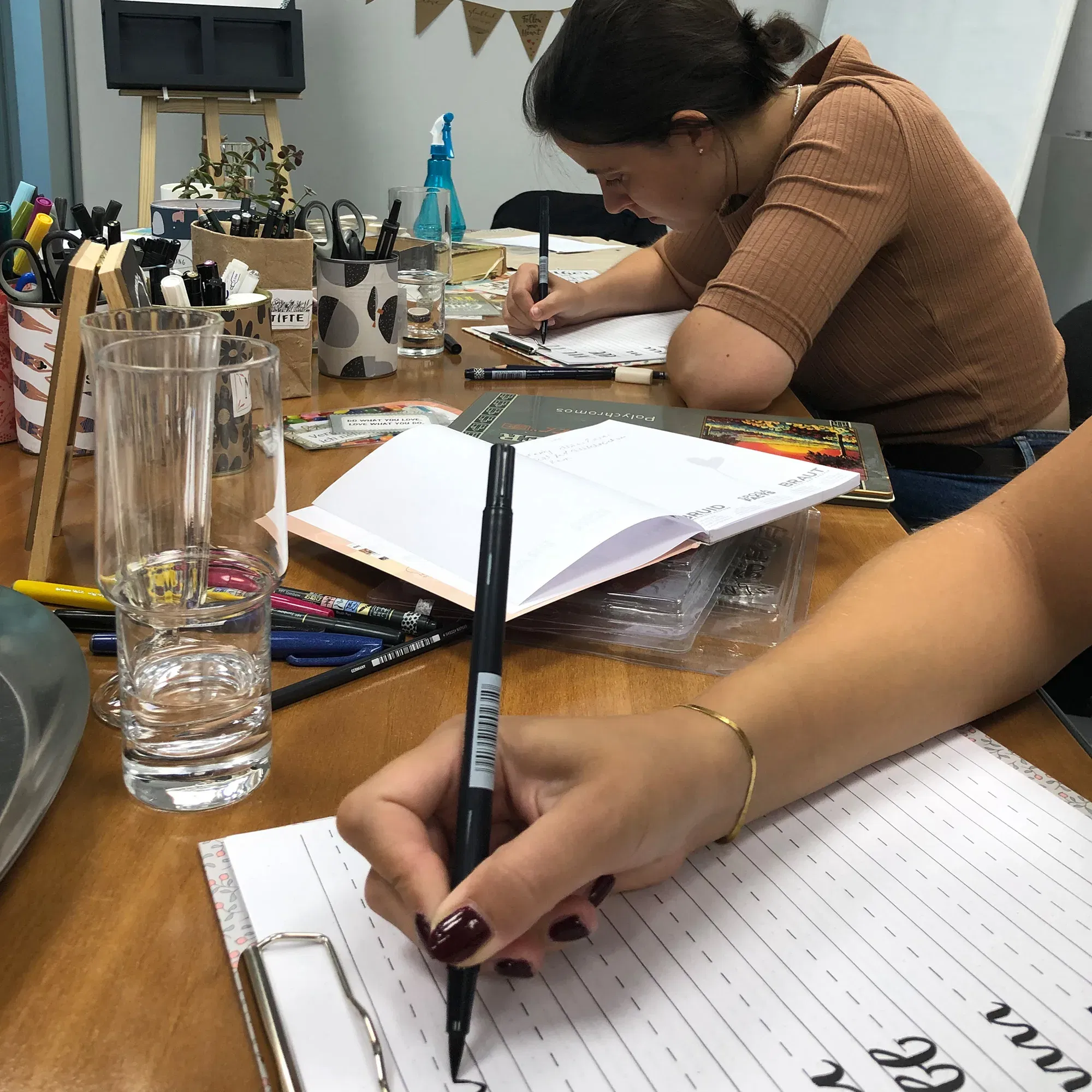 Eine Workshop-Szene zeigt Menschen, die Handlettering-Übungen an einem gut ausgestatteten Tisch machen. Stifte, Notizblöcke und Gläser sind über den Tisch verteilt, während eine Teilnehmerin eifrig schreibt.