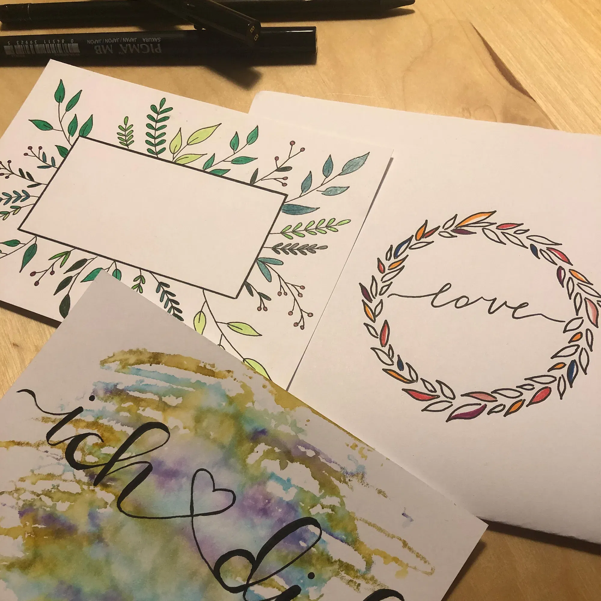 Drei handgemachte Karten mit kunstvollen Handlettering-Designs liegen auf einem Tisch. Eine Karte zeigt den Schriftzug „ich liebe dich“ in Aquarell, die andere „love“ in einem Kranz aus Blättern.
