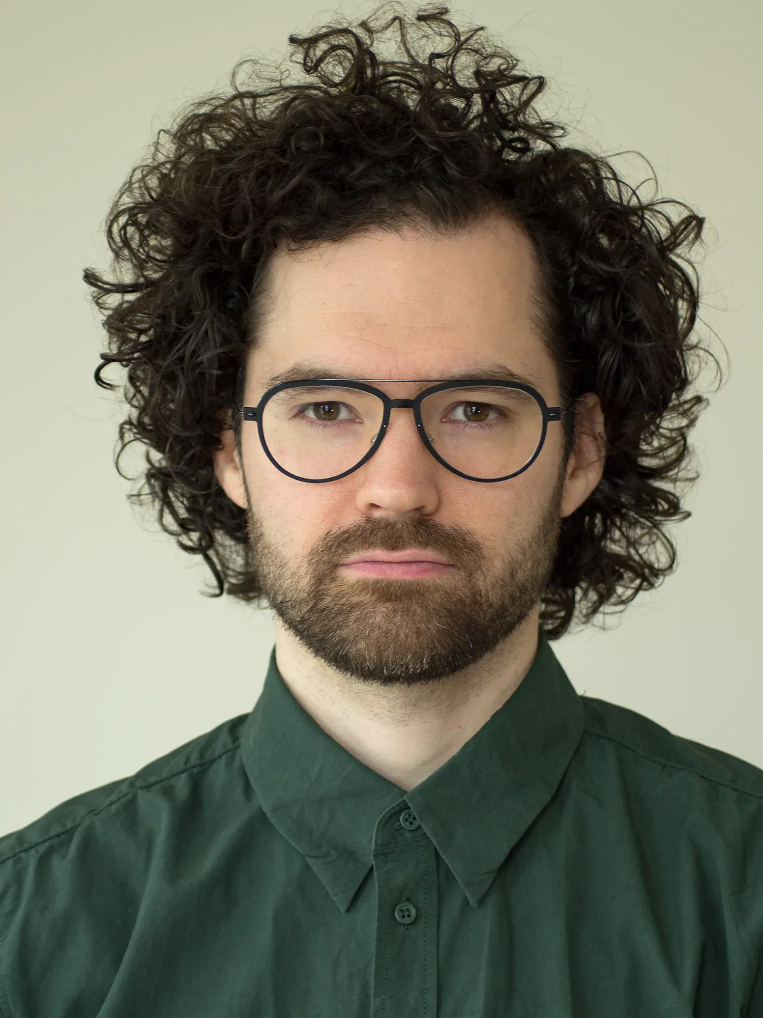 Porträt von Lukas Grewenig, der mit Brille und lockigem Haar im grünen Hemd vor einem hellen Hintergrund ernst schaut.