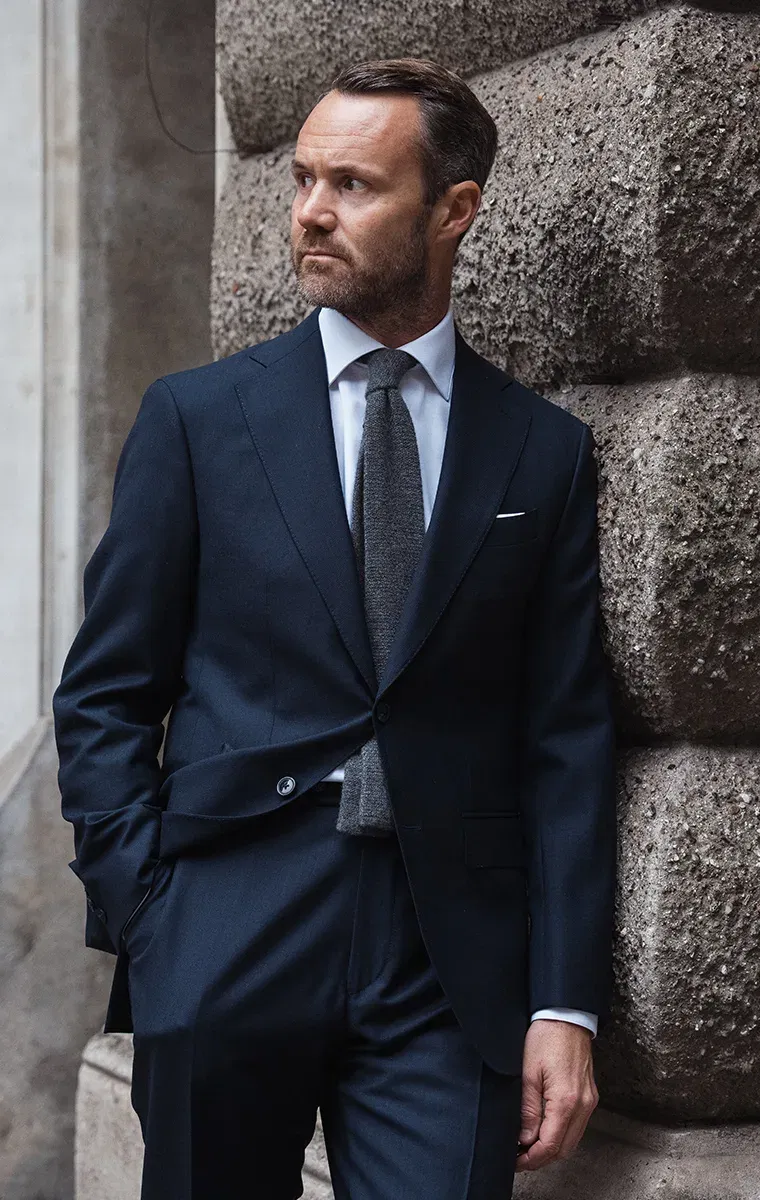 Elegant gekleideter Mann in dunklem Businessanzug lehnt an einer Steinwand und blickt zur Seite.