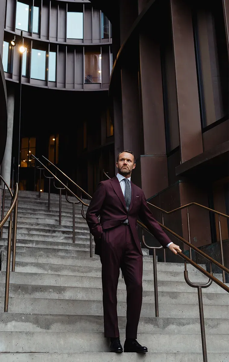 Mann im bordeauxfarbenen Anzug steht auf einer modernen Treppe in urbaner Architektur und schaut in die Ferne.
