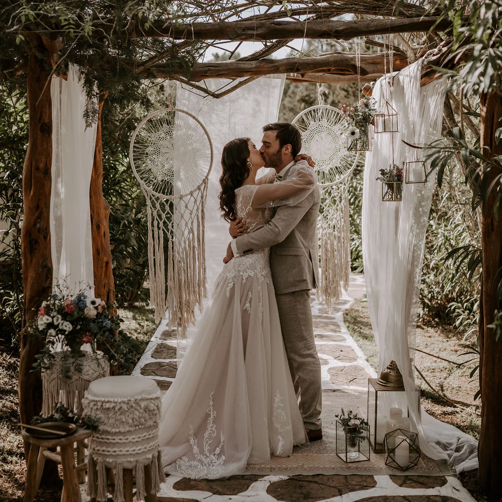 Romantischer Kuss des Brautpaars bei einer Boho-Hochzeit unter einem von Traumfängern und Makramees verzierten Traubogen.