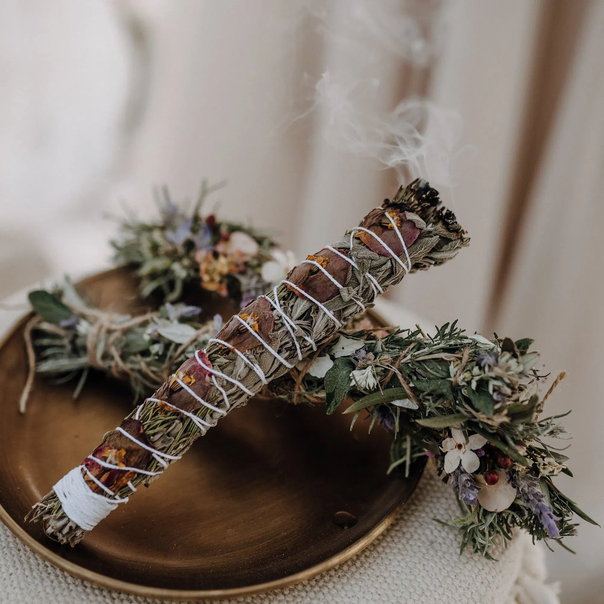 Räucherbündel aus Kräutern und Blüten auf einem dekorativen Teller, brennend und umgeben von natürlichen Blumenarrangements.
