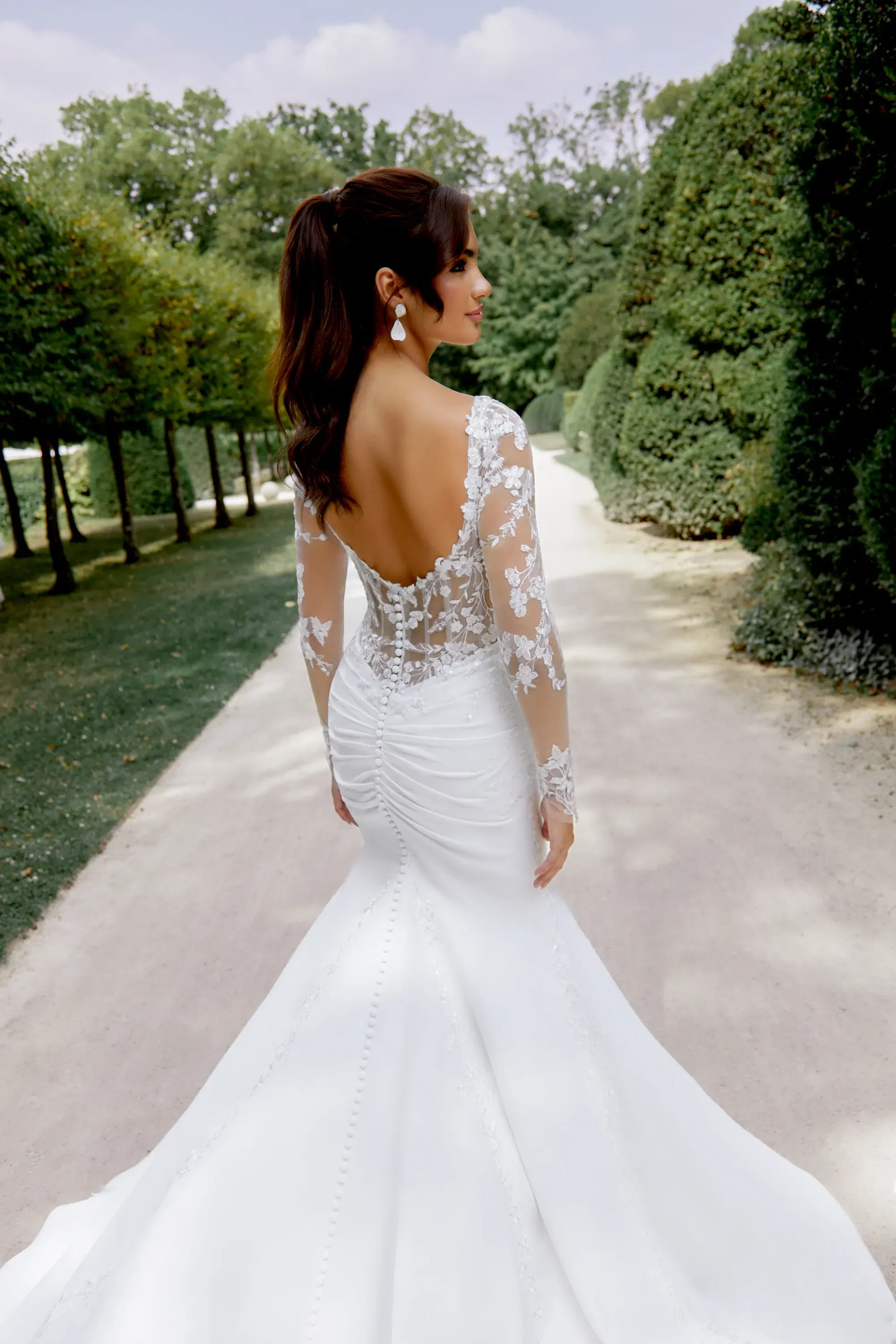 Rückansicht eines figurbetonten Brautkleids mit transparenter Spitzenrückenpartie und Knopfleiste – aufgenommen im Park.