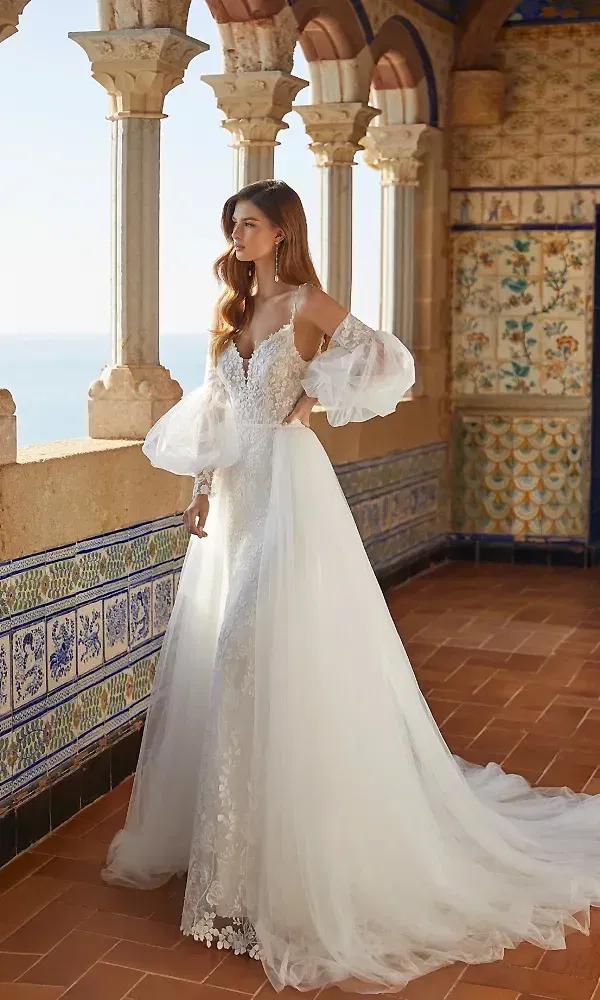 Die Braut trägt ein fließendes, bodenlanges Kleid mit transparenten Ballonärmeln und zarter Spitze, umgeben von kunstvollen Fliesen und Arkaden auf einem mediterranen Balkon. Der Meerblick und die architektonischen Details im Hintergrund verleihen der Szene eine verträumte, majestätische Atmosphäre.