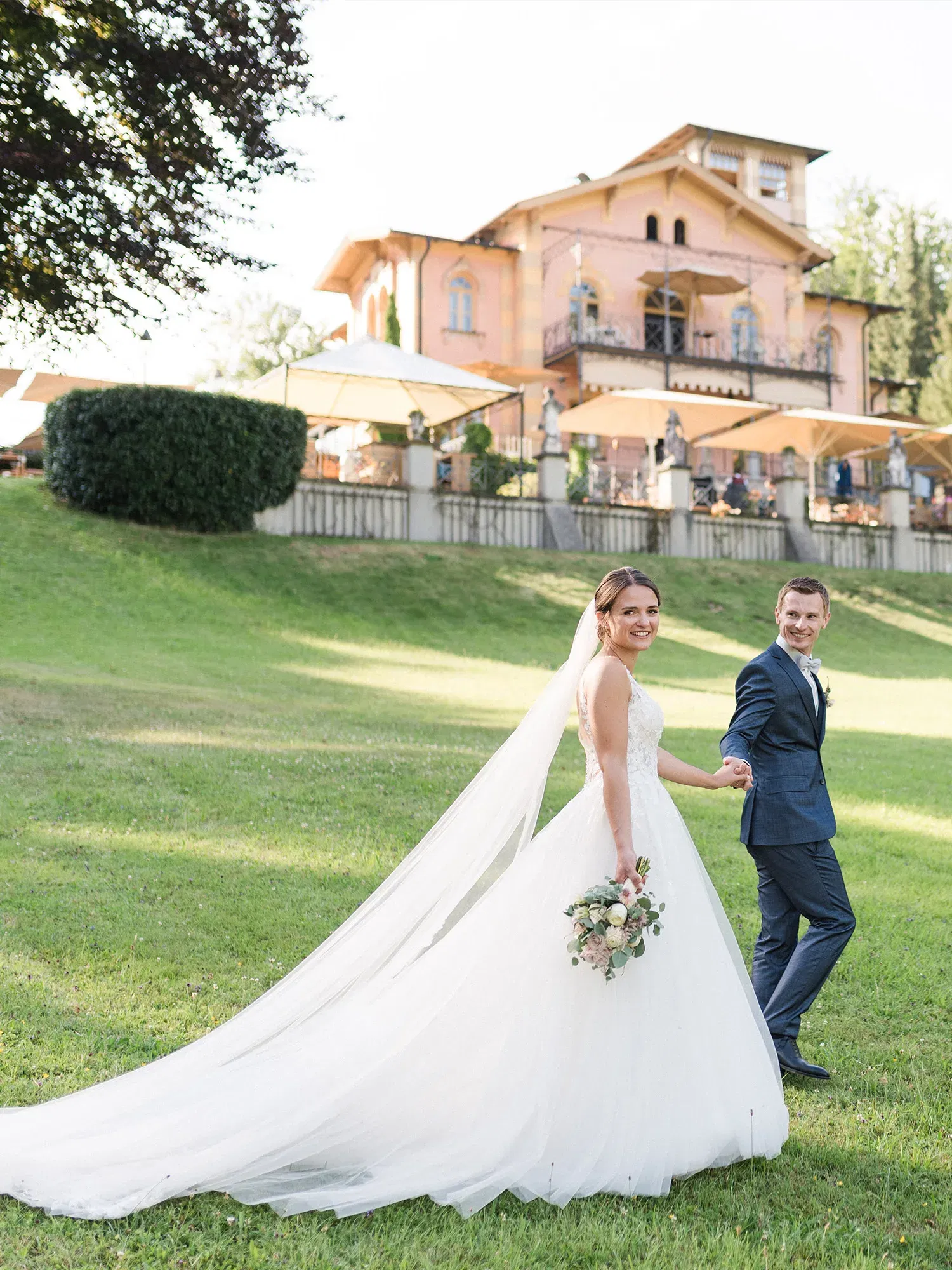 Eine Braut und ihr Bräutigam laufen lächelnd auf einem weitläufigen Rasen. Die Villa im Hintergrund und die sanft fließende Schleppe der Braut verleihen der Szene eine märchenhafte Atmosphäre.