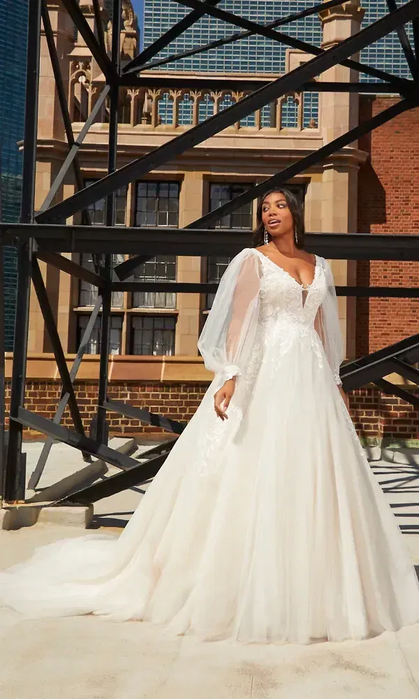 Vor einem imposanten Gebäude mit Glasfassade steht eine Braut in einem eleganten A-Linien-Hochzeitskleid mit transparenten Ärmeln und tiefem Ausschnitt. Der Kontrast zwischen der modernen Architektur und dem klassischen Kleid ist auffällig.