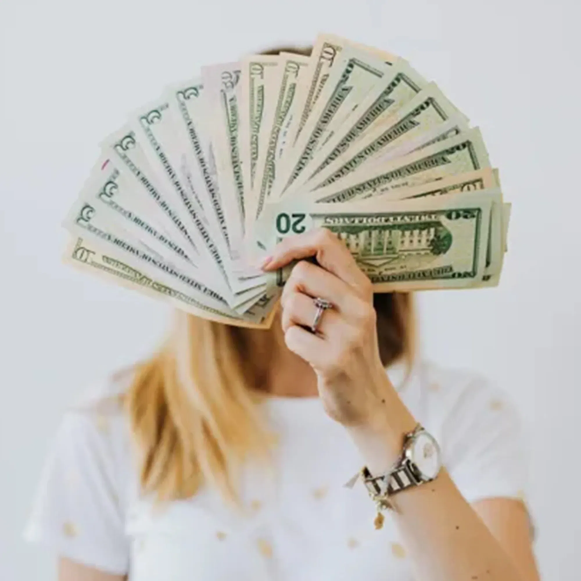 Das Bild zeigt eine Frau, die einen Fächer aus US-Dollar-Scheinen hochhält, wobei ihr Gesicht hinter den Geldscheinen verborgen ist. Sie trägt ein weißes Oberteil und eine auffällige Armbanduhr.