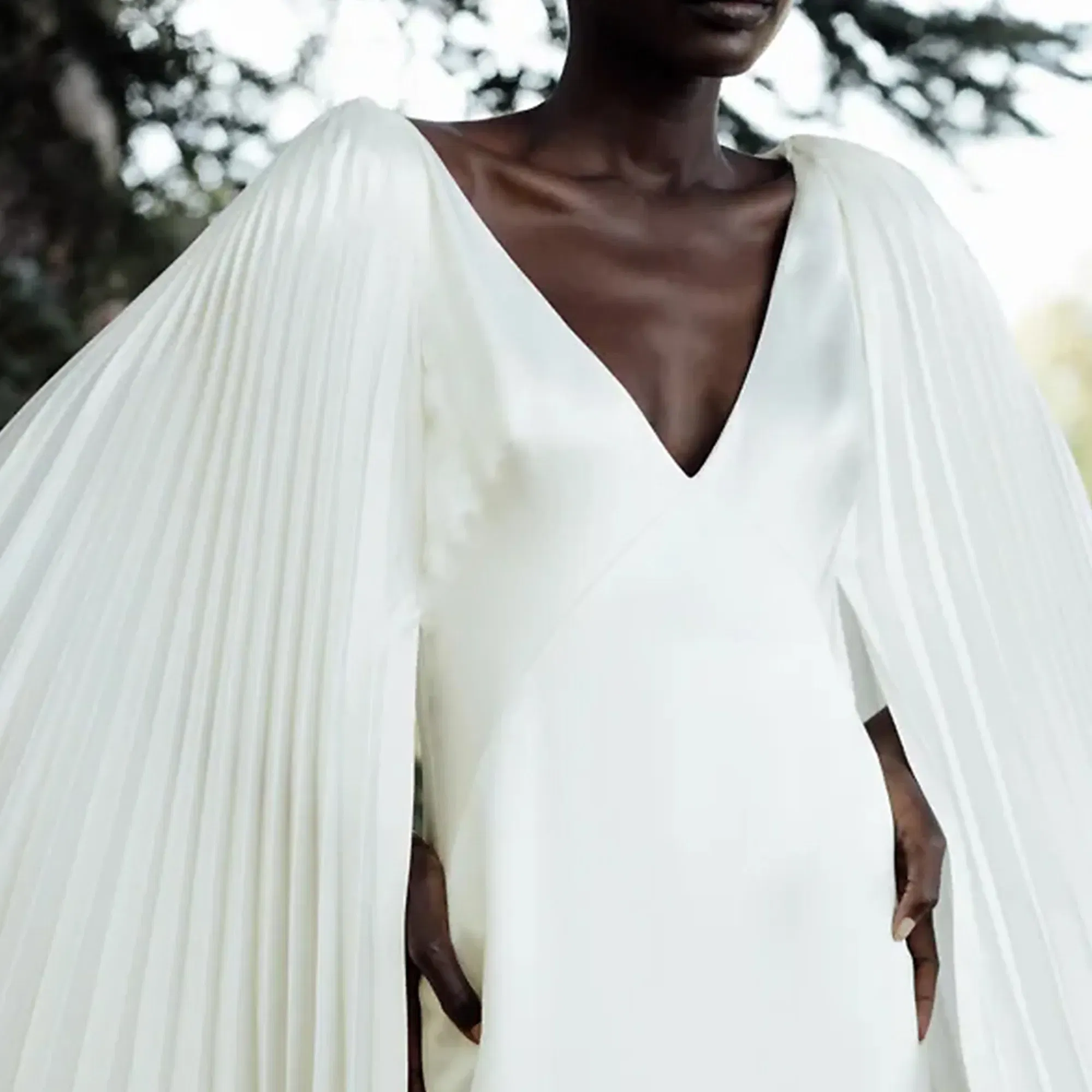 Ein Brautkleid mit tiefem V-Ausschnitt und auffälligen, plissierten Ärmeln, das einen modernen und zugleich zeitlosen Look vermittelt. Die Braut steht im Freien und das Kleid hebt sich kontrastreich vor dem Hintergrund ab.