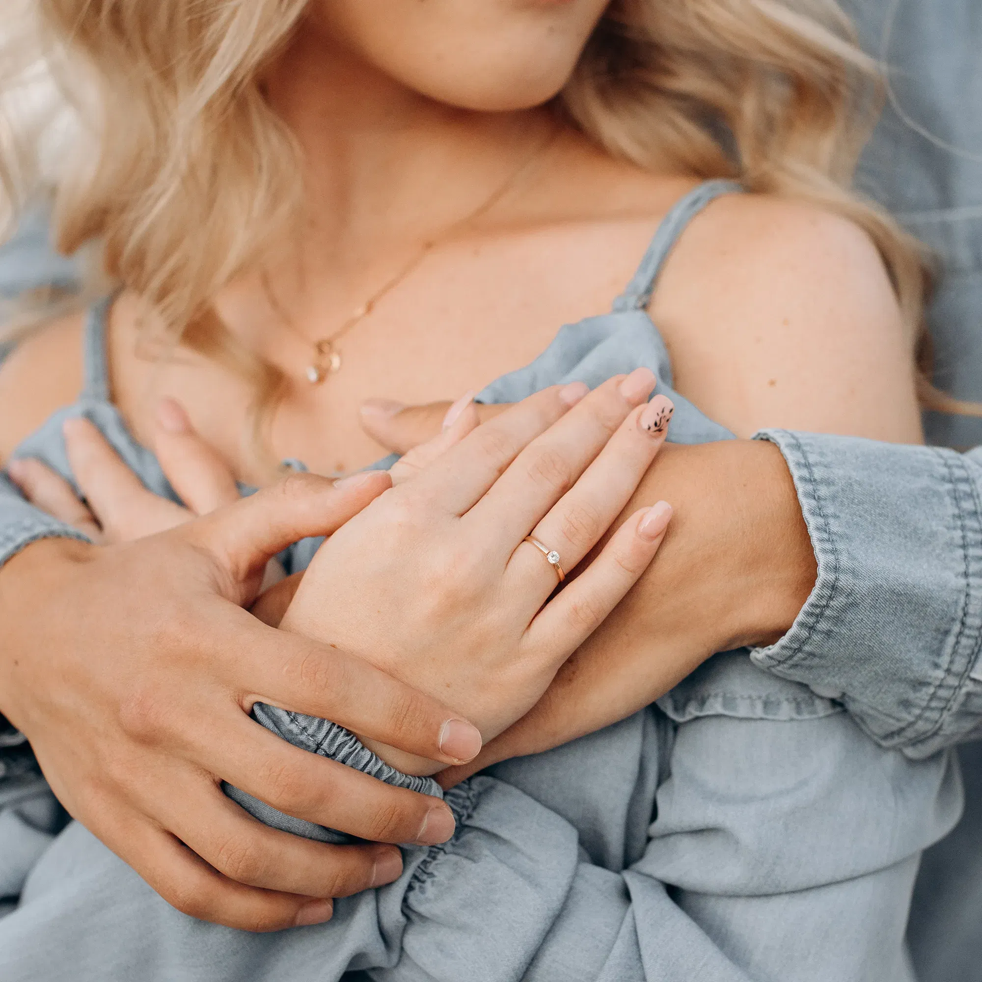 Eine Frau hält die Hände ihres Partners liebevoll an ihrer Schulter, wobei ihr zarter Verlobungsring im Vordergrund glänzt.