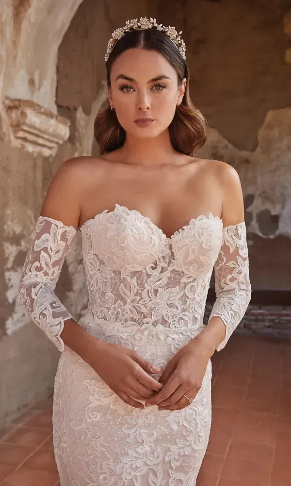 In einem wunderschönen, trägerlosen Brautkleid mit zarten Spitzenapplikationen und transparenten Ärmeln steht die Braut vor einer antiken Steinwand. Ihr Kopfschmuck aus funkelnden Perlen und Blüten ergänzt das romantische, stilvolle Gesamtbild.