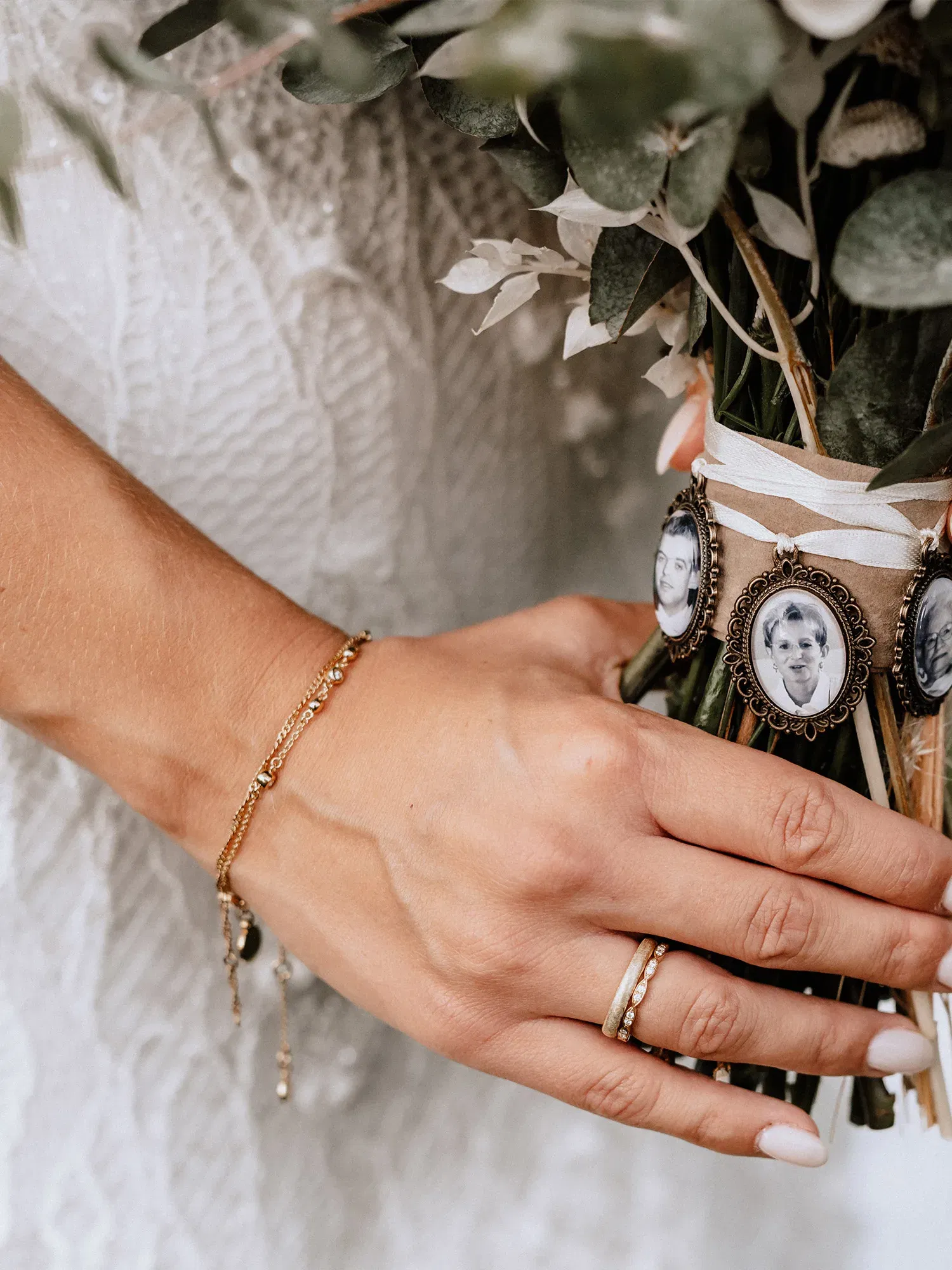 Die Hand einer Braut, die sanft einen Blumenstrauß hält, verziert mit gerahmten Porträtbildern von Familienmitgliedern. Ihre Hand trägt einen Ehering und ein filigranes Armband.