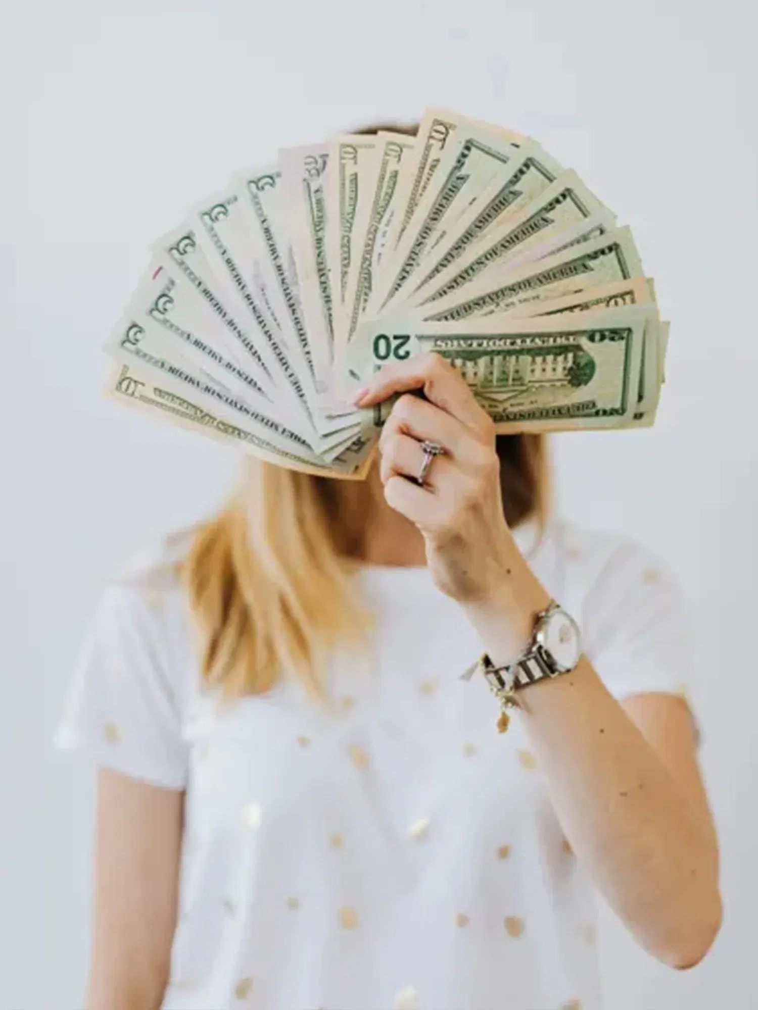 In diesem Bild hält eine Frau mehrere Dollar-Scheine wie einen Fächer vor ihr Gesicht, wobei ihr Arm und eine Armbanduhr klar sichtbar sind. Das weiße T-Shirt mit goldenen Punkten hebt sich vom neutralen Hintergrund ab.