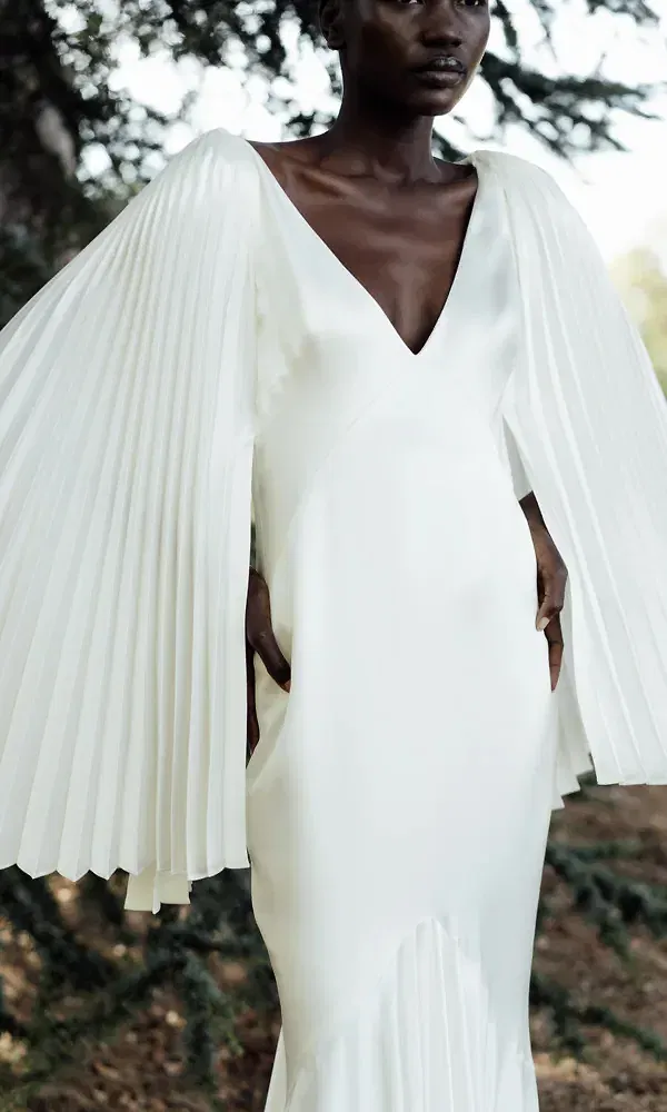 Eine Braut in einem avantgardistischen Brautkleid, das durch voluminöse, plissierte Ärmel und einen tiefen V-Ausschnitt besticht. Die Struktur des Kleides verleiht dem Look eine Mischung aus Eleganz und künstlerischem Flair, passend für eine moderne Braut.