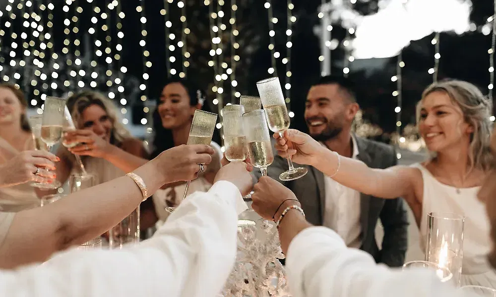 Gäste einer abendlichen Hochzeit stoßen mit Sektgläsern an. Im Hintergrund sorgen funkelnde Lichterketten für eine stimmungsvolle Kulisse. Die fröhlichen Gesichter der Gäste unterstreichen die ausgelassene Feierlaune.
