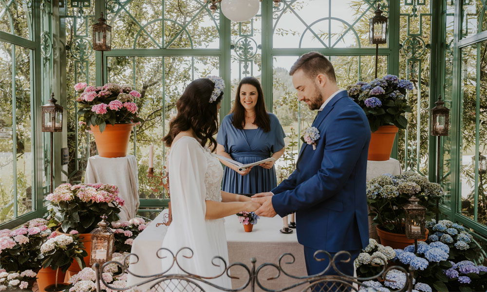 Eine romantische freie Trauung in einem Gewächshaus, umgeben von Hortensien. Das Brautpaar tauscht Blicke, während die Traurednerin lächelt und die Zeremonie führt.