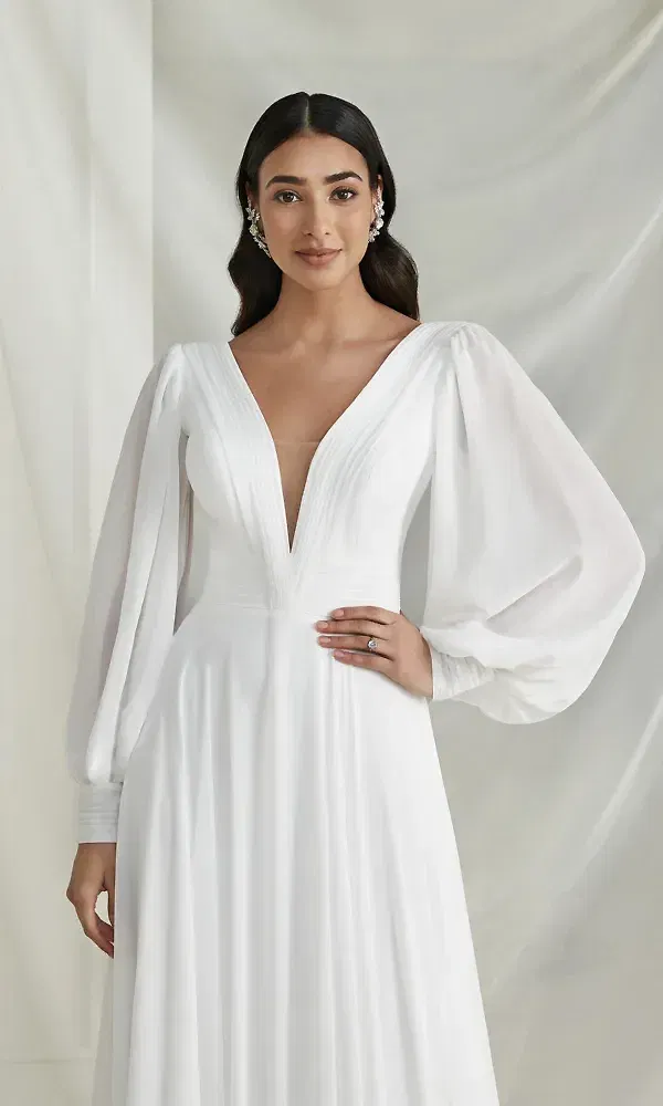 Eine stilvolle Braut steht in einem klassischen, weißen Brautkleid mit langen, weiten Ärmeln und tiefem Ausschnitt. Das Kleid zeichnet sich durch schlichte Raffinesse aus, mit fließendem Stoff, der den femininen Look unterstreicht. Ihr dezent geschmückter Haarschmuck vervollständigt das Gesamtbild.