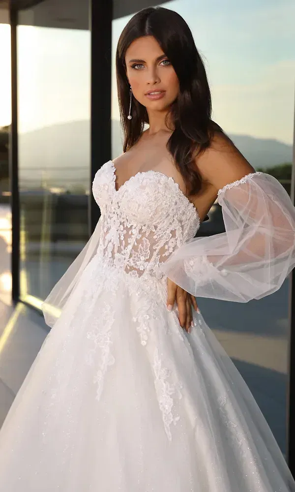 In einem luxuriösen, trägerlosen Brautkleid mit filigranen Spitzenapplikationen und transparenten Tüllärmeln steht die Braut vor einer modernen Glaswand. Das Licht der untergehenden Sonne reflektiert auf dem glitzernden Rock des Kleides, was den gesamten Look märchenhaft und glamourös erscheinen lässt.