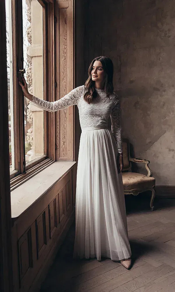 Das Bild zeigt eine Braut in einem Vintage-inspirierten Hochzeitskleid mit langen Spitzenärmeln. Sie steht verträumt am Fenster, ihr Kleid fällt in sanften Falten zu Boden, während die rustikale Umgebung dem Bild eine nostalgische Atmosphäre verleiht.