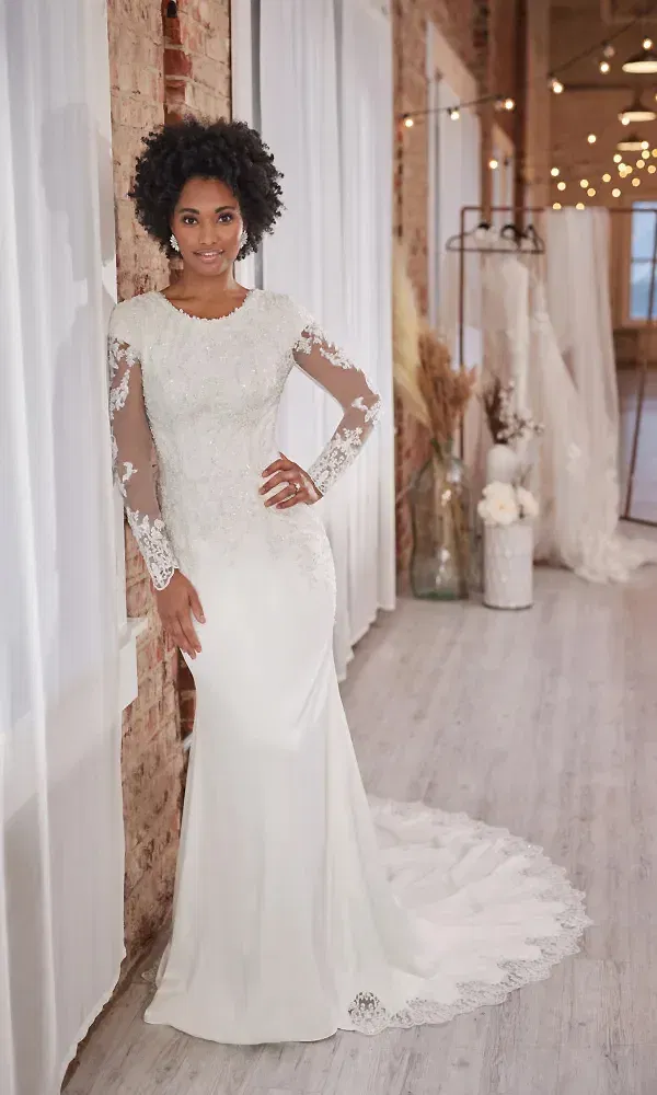 Eine Braut in einem detailreich gestalteten Hochzeitskleid mit zarten Spitzenapplikationen an den Ärmeln und einer langen Schleppe lehnt sich gegen eine Backsteinwand. Die Umgebung ist festlich beleuchtet und mit sanften Tönen dekoriert, passend zum edlen Stil des Kleides.