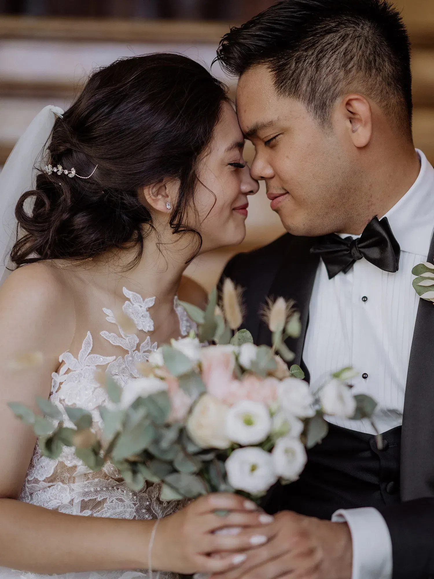 Ein Brautpaar steht eng zusammen, ihre Stirnen berühren sich, während sie lächeln. Die Braut hält einen Strauß aus weißen und zartrosafarbenen Blumen, während der Bräutigam einen klassischen schwarzen Anzug mit Fliege trägt.