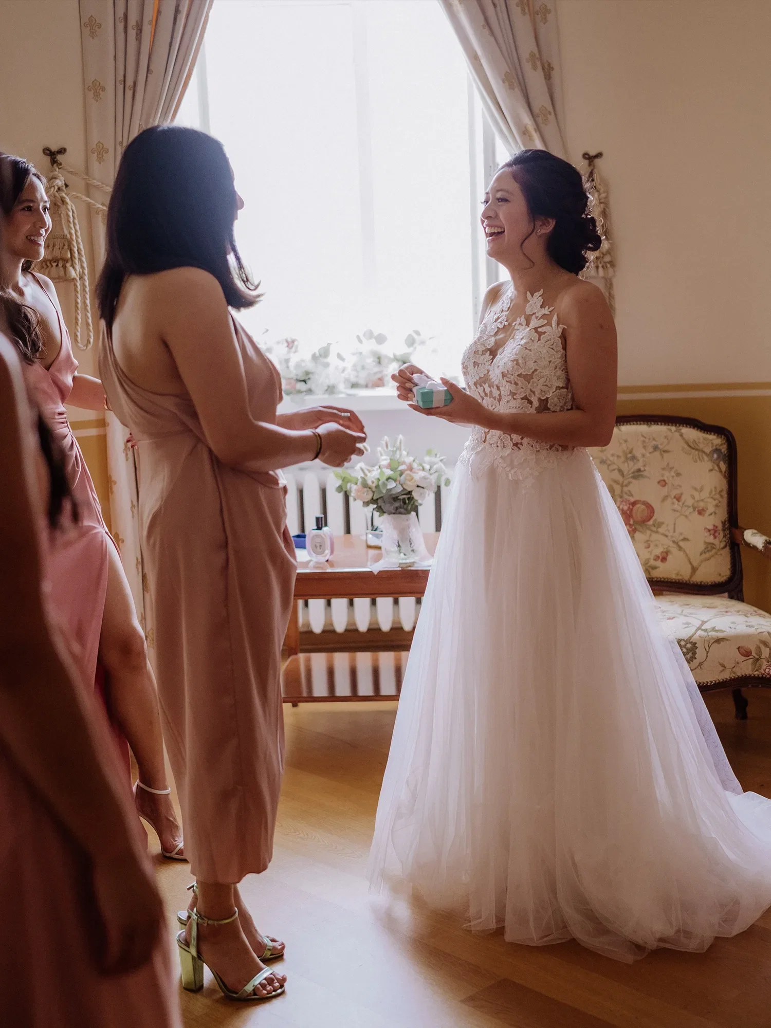 Die Braut steht in einem lichtdurchfluteten Zimmer und lächelt, während sie ein Geschenk von einer ihrer Brautjungfern entgegennimmt. Beide tragen festliche Kleidung, und die Szene strahlt Freude und Vorfreude aus.