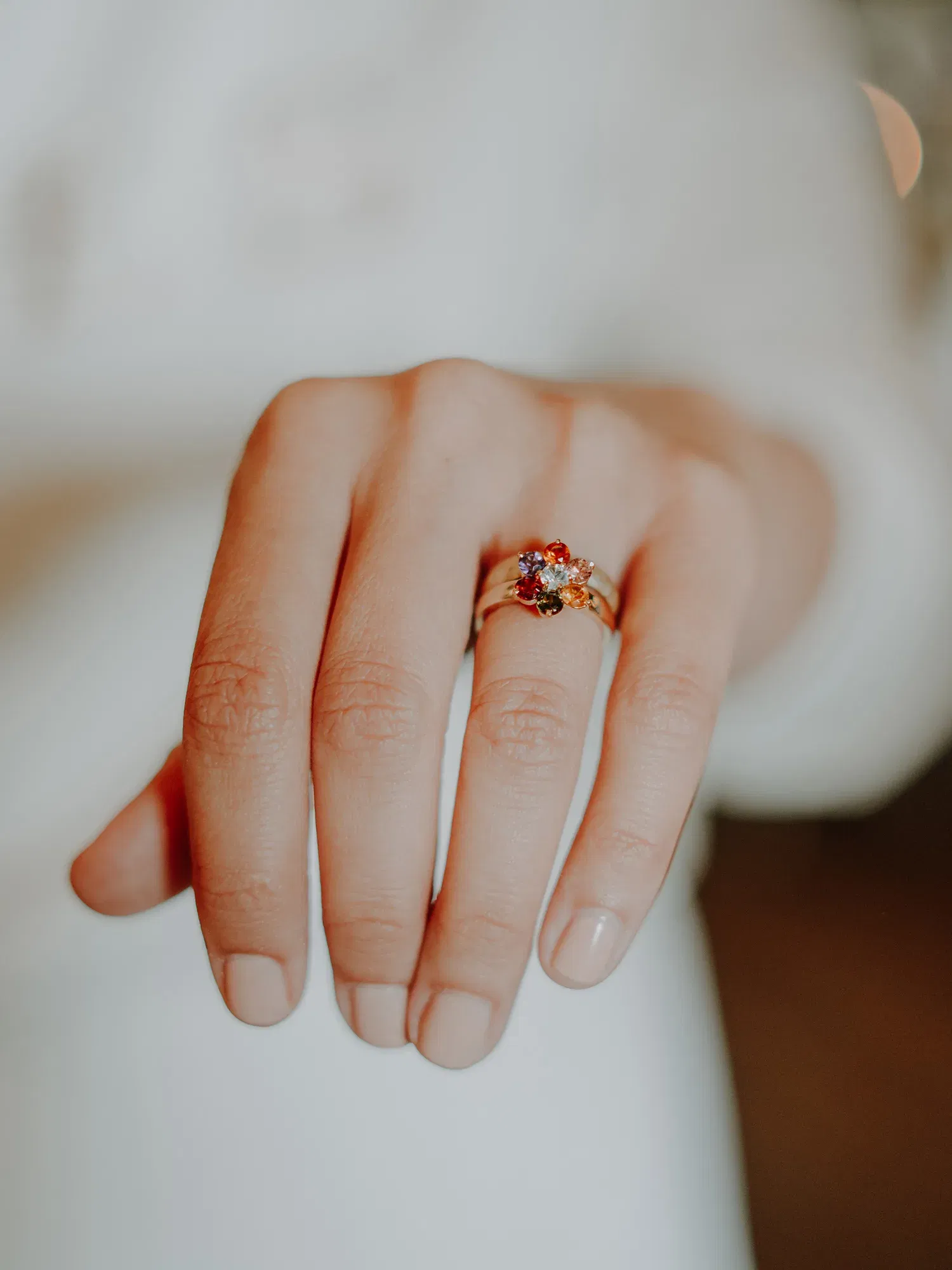 Im Zentrum des Bildes steht eine elegante Hand mit einem auffälligen Ring, dessen Edelstein in kräftigen Farben funkelt. Der Fokus liegt auf dem Schmuckstück, das vor einem verschwommenen, warmen Hintergrund in Szene gesetzt wird.